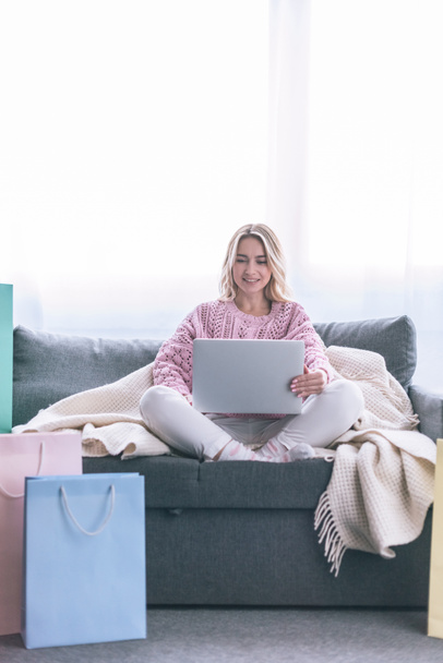 happy blonde woman sitting on sofa with laptop near shopping bags - Foto, imagen