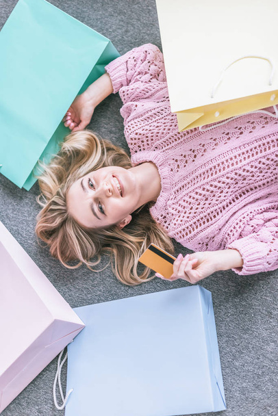  top view of cheerful woman holding credit card and lying on floor near shopping bags - Photo, Image
