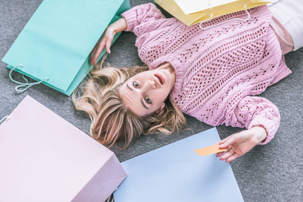  top view of surprised woman holding credit card and lying on floor near shopping bags - Fotoğraf, Görsel