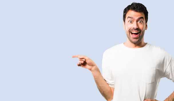 Jovem com camisa branca apontando o dedo para o lado e apresentando um produto enquanto sorri em uma pose confiante no fundo azul isolado
 - Foto, Imagem