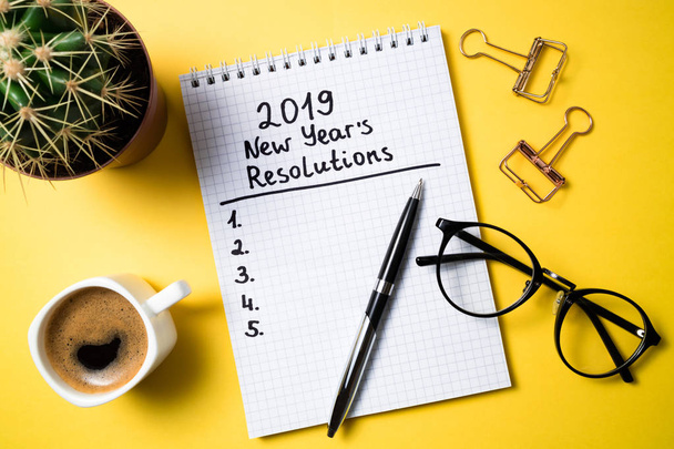 Resolución de año nuevo 2019 en el escritorio. Lista de goles 2019 con cuaderno, taza de café y vasos sobre fondo amarillo. Vista superior
 - Foto, Imagen