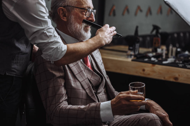 Bearded old businessman sitting at barber shop in chair choosing haircut design - Photo, Image