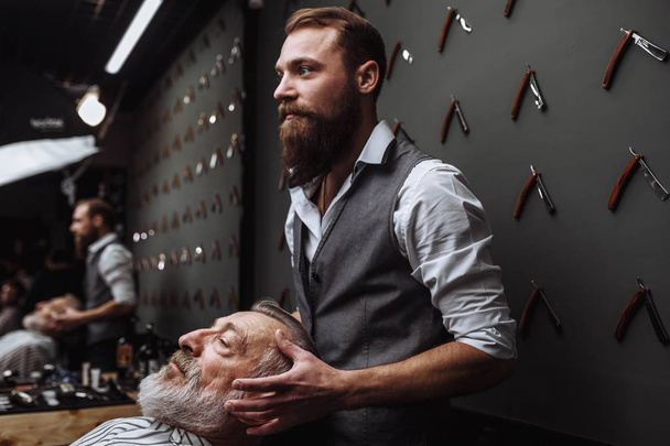 Male barber chooses beard design for old-aged client at hairdresser. - 写真・画像