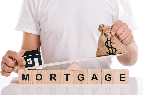 cropped view of man holding moneybag and paper house on scales on cubes with mortgage lettering isolated on white - Photo, Image