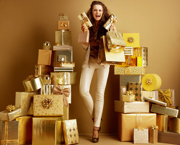 Full length portrait of happy stylish woman in gold beige clothes with gifts and shopping bags among 2 piles of golden gifts in front of a plain wall. concept of abundance of gifts for everyone. - Фото, зображення