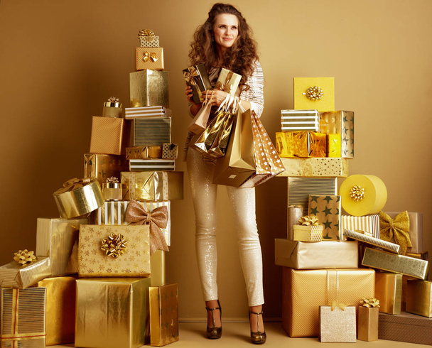 Full length portrait of pensive elegant woman in gold beige clothes with gifts and shopping bags looking into the distance among 2 piles of golden gifts in front of a plain wall. holidays shopping - Fotografie, Obrázek