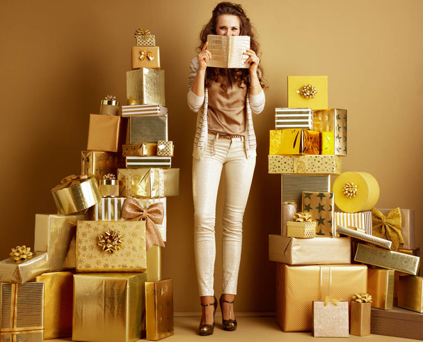 Full length portrait of smiling elegant fashion-monger in gold beige clothes among 2 piles of golden gifts in front of a plain wall holding notebook in the front of face. making list of holiday gifts - Valokuva, kuva