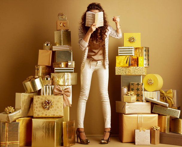 Full length portrait of trendy fashion-monger in gold beige clothes among 2 piles of golden gifts in front of a plain wall holding diary in the front of face. making list of gift ideas - Valokuva, kuva