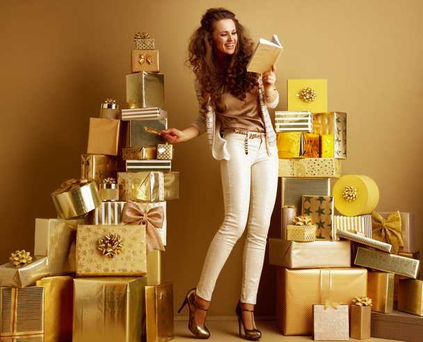 Full length portrait of smiling stylish fashion-monger in beige clothes looking at notebook and counting gift boxes, holidays shopping concept  - Фото, изображение
