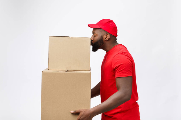 Conceito de entrega - Vista lateral Retrato de homem de entrega afro-americano feliz em pano vermelho segurando um pacote de caixa. Isolado no fundo cinzento. Espaço de cópia
 - Foto, Imagem
