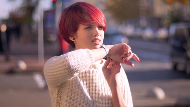 Ritratto di felice bella ragazza dai capelli rosa chiamando utilizzando il suo orologio su sfondo strada offuscata
. - Filmati, video