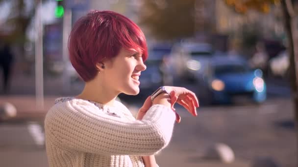 Retrato de chica de pelo rosa bonita emocional llamando usando su reloj en el fondo borroso de la calle
. - Metraje, vídeo