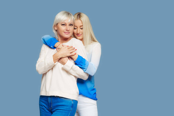 Teen blond daughter and mature mother on blue background hugging - Photo, image