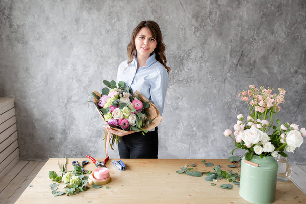 Young friendly smiling female floral artist looking at the camera, holds a luxurious bouquet in his hands. Professional florist making a bouquet of pink Ranunculus and peonies in a flower shop. - Fotoğraf, Görsel