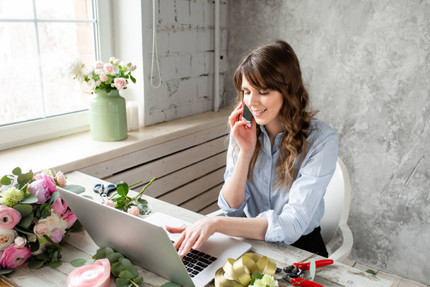 Young friendly smiling female florist talking on a mobile phone and create ordering in the working base on a laptop. Flower delivery concept. Professional worker in a flower shop. - Foto, imagen