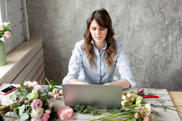 junge freundlich lächelnde Floristin schafft Ordnung in der Arbeitsbasis auf einem Laptop. Blumenlieferkonzept. Facharbeiter in einem Blumenladen. - Foto, Bild