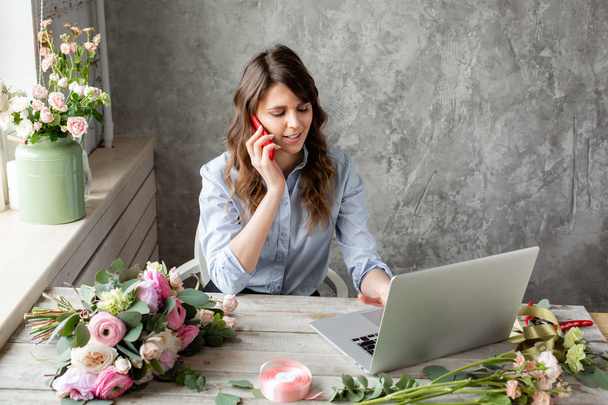 Young friendly smiling female florist talking on a mobile phone and create ordering in the working base on a laptop. Flower delivery concept. Professional worker in a flower shop. - Foto, afbeelding