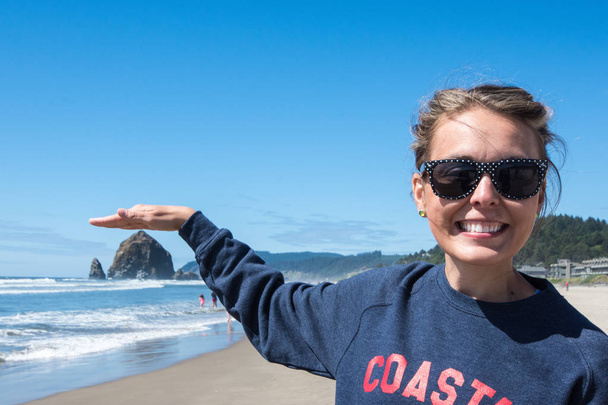 La donna che indossa gli occhiali da sole finge di toccare una roccia Haystack a Cannon Beach, Oregon sulla spiaggia in una giornata di sole. Vista prospettiva forzata
 - Foto, immagini