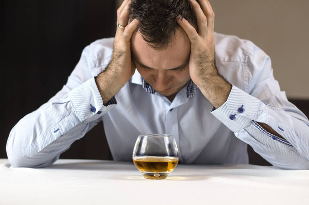 アルコールのトラブル。落ち込んでいる男は、彼の手で彼の頭をテーブルに座る。ウィスキーのグラスが彼の前に立っています。. - 写真・画像