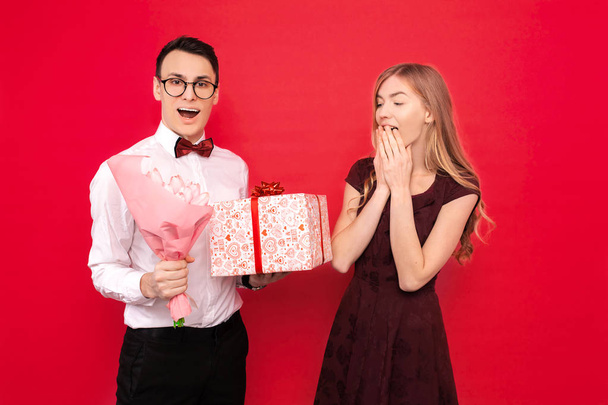 A handsome student, wearing glasses, holding books in her hands, gives a gift and a bouquet of flowers to his girlfriend against a red background. Valentine's Day - Photo, image