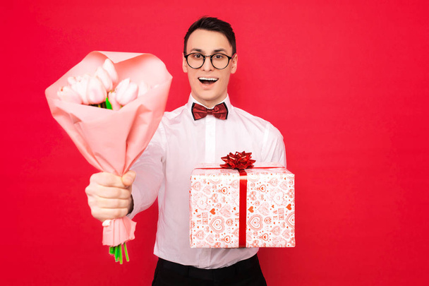 man, wearing glasses, with a gift and a bouquet of flowers, on a red background, the concept of women's day - 写真・画像