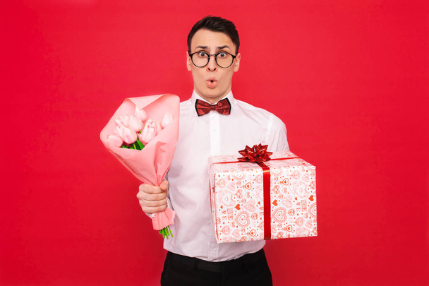 Surprised man in glasses, with a gift and a bouquet of flowers, on a red background - Foto, immagini