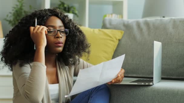 Portrait shot of tired young African American woman in glasses working hard with chart document and laptop in the living room. - Imágenes, Vídeo
