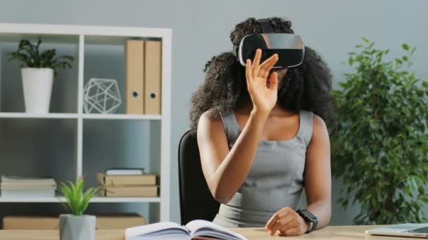 Pretty curly African American young woman using virtual reality headset, VR glasses while sitting at the table in the office. Indoor - Кадры, видео