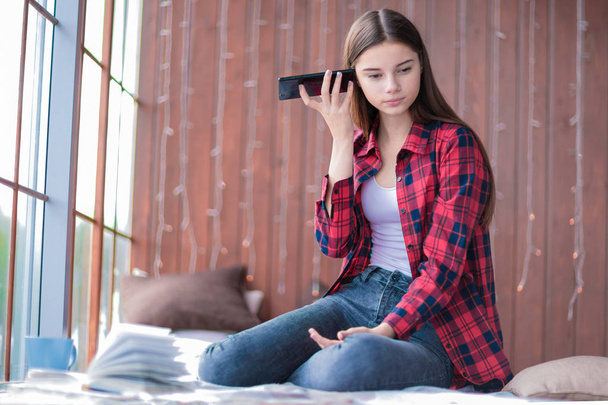 Симпатичная девушка слушает голосовое сообщение своего мобильного телефона, сидящего на полу у окна
. - Фото, изображение