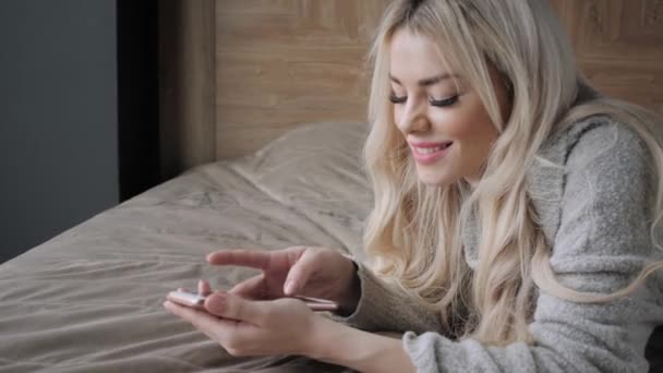 Beautiful blonde woman lying on the bed with smart phone. Girl smiles, good mood. Blogging, browsing internet, chatting. In a warm cozy sweater and wool socks. Happy winter concept. - Video