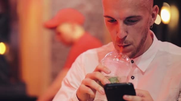 Young Attractive Man Using Smartphone at Evening. He sitting in a bar or restaurant near neon signage and drinking cocktail. Communication, rest, chatting, travel concept. Close up - Кадри, відео