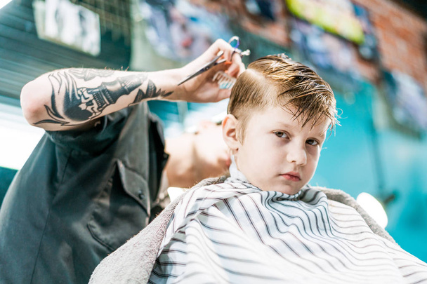 A boy is looking in camera while being cut his hairstyle in barbershop. - Фото, зображення