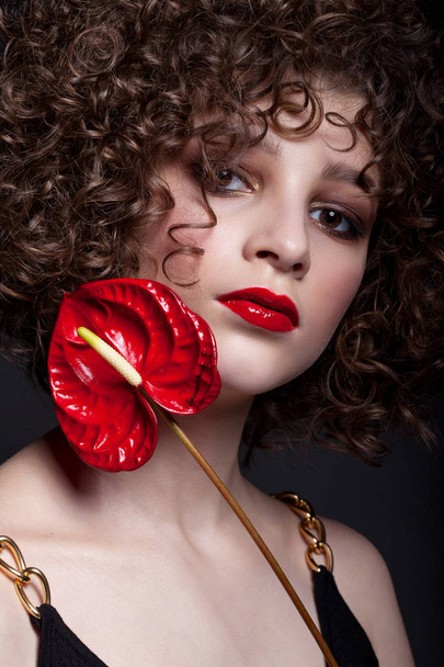 Portrait d'une belle adolescente avec un maquillage lumineux. Avec des boucles afro et rouge à lèvres rouge et une fleur rouge sur le visage. Sur fond noir
. - Photo, image