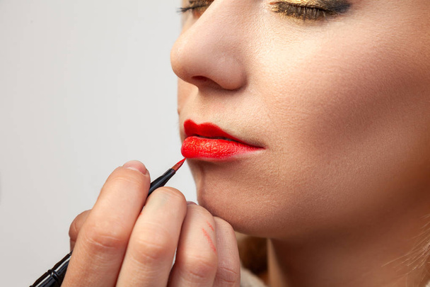 Close-up of make-up applying on the model's lips, the make-up artist holds a brush in her hand and applies red lipstick, the blonde girl's eyes are closed, oriental-style eye shadows on her eyelids. - Photo, Image
