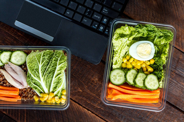 Healthy, dietary meal in a plastic container and a laptop on a wooden background. Snack in the office, at work. Lunch box. Nutrition concept. Food on schedule, lunch break - Foto, imagen
