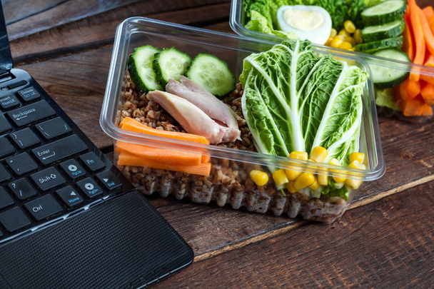 Healthy, dietary meal in a plastic container and a laptop on a wooden background. Snack in the office, at work. Lunch box. Nutrition concept. Food on schedule, lunch break - Fotoğraf, Görsel