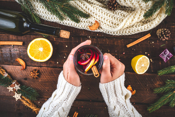 A young woman in a warm, knitted, white pullover is holding a Christmas, hot mulled wine in a glass with spices and citrus fruits. A cozy, winter evening. Winter drinks. Flat lay, top view  - Φωτογραφία, εικόνα
