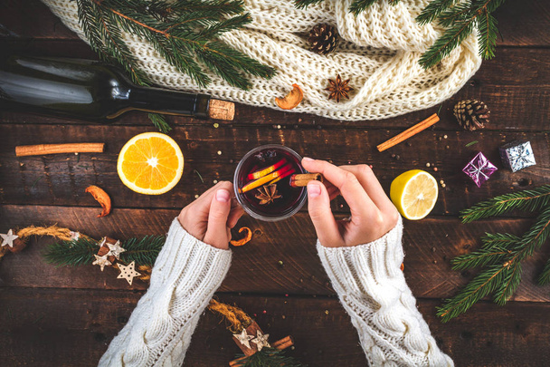 A young woman in a warm, knitted, white pullover is holding a Christmas, hot mulled wine in a glass with spices and citrus fruits. A cozy, winter evening. Winter drinks. Flat lay, top view  - Photo, Image