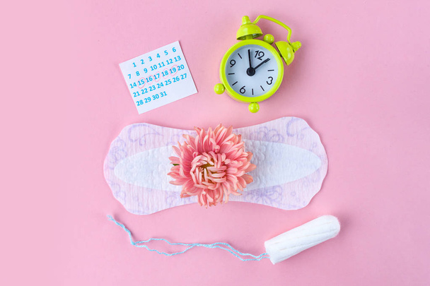 Tampons for menstruation, alarm clock, feminine pads and a pink flower on a pink background. Hygiene care during critical days. Regular menstrual cycle.  - Photo, Image
