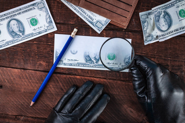 Counterfeiter in black gloves forges banknotes. Fake concept. Fake money, a wallet with American dollars, a magnifier on a wooden background. Top view  - Zdjęcie, obraz