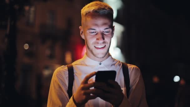Νεαρός άνδρας ελκυστική χρησιμοποιώντας smartphone στέκεται στον δρόμο της πόλης το βράδυ. Επικοινωνία, ταξίδια, τουρισμός, χρονολόγηση, επιχειρηματική ιδέα. - Πλάνα, βίντεο