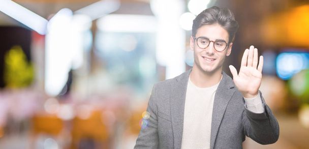 Young business man wearing glasses over isolated background Waiving saying hello happy and smiling, friendly welcome gesture - Photo, Image