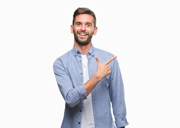 Jeune bel homme portant un t-shirt blanc sur fond isolé gai avec un sourire du visage pointant avec la main et le doigt sur le côté avec une expression heureuse et naturelle sur le visage regardant la caméra
. - Photo, image