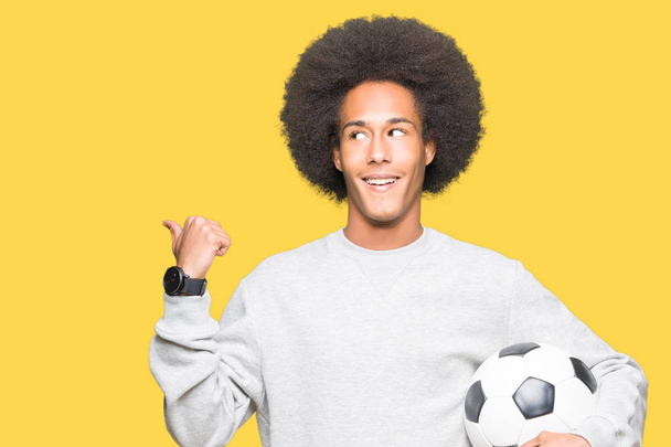 Young african american man with afro hair holding soccer football ball pointing and showing with thumb up to the side with happy face smiling - Fotó, kép