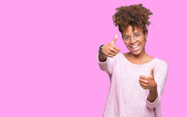 Beautiful young african american woman wearing glasses over isolated background approving doing positive gesture with hand, thumbs up smiling and happy for success. Looking at the camera, winner gesture. - Photo, Image