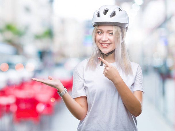 Молода блондинка в шоломі велосипедиста над ізольованим тлом здивована і посміхається камері, представляючи рукою і вказуючи пальцем
. - Фото, зображення