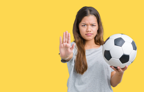Молодая красивая брюнетка женщина держит футбольный мяч на изолированном фоне с открытой рукой делает знак стоп с серьезным и уверенным выражением, защитный жест
 - Фото, изображение