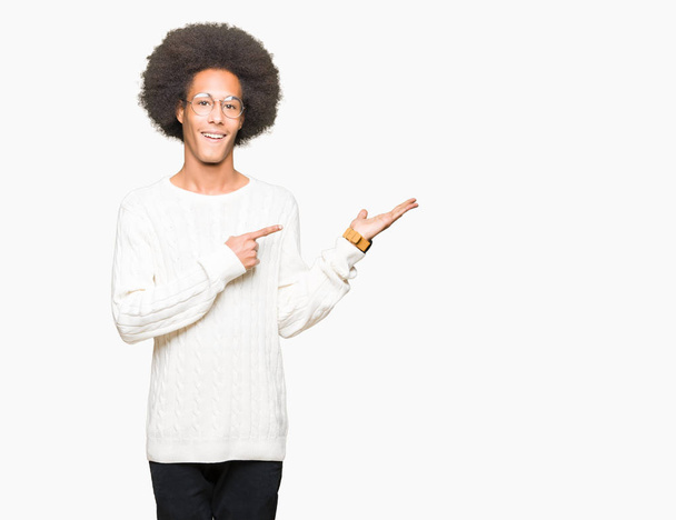 Jonge Afro-Amerikaanse man met afro haar bril verbaasd en lachend naar de camera tijdens het presenteren met hand en met de vinger te wijzen. - Foto, afbeelding