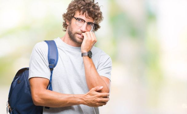 Όμορφος Ισπανόφωνος φοιτητής άνδρα που φοράει γυαλιά και σακίδιο πέρα από το απομονωμένο υπόβαθρο ψάχνει αγχωμένοι και νευρικοί με τα χέρια στο στόμα δάγκωμα νύχια. Πρόβλημα του άγχους. - Φωτογραφία, εικόνα