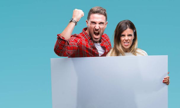 Young couple together holding blank banner over isolated background annoyed and frustrated shouting with anger, crazy and yelling with raised hand, anger concept - Photo, Image